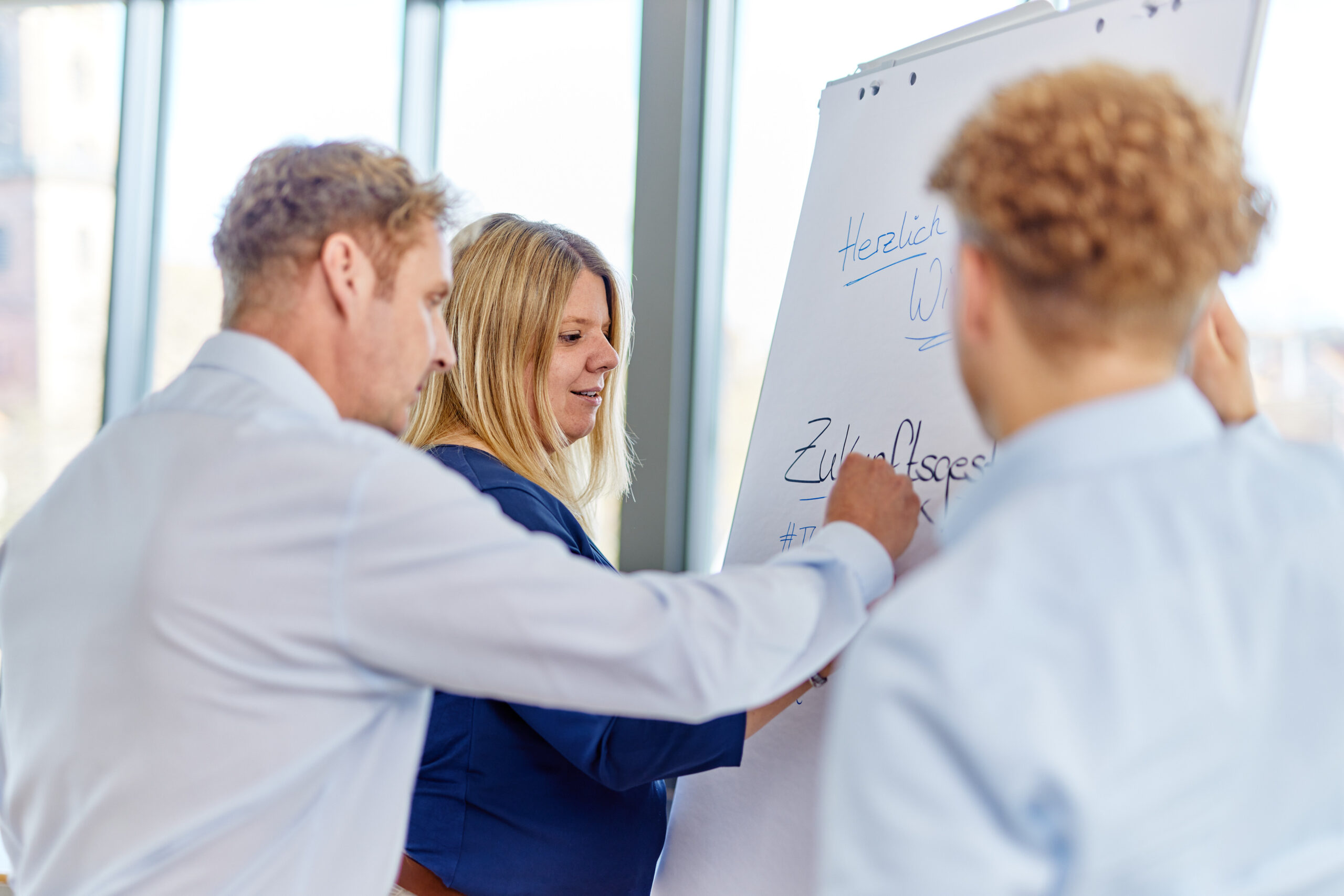 Drei Personen stehen vor einem Flipchart in einem modernen Besprechungsraum mit großen Fenstern; eine Frau hält das Flipchart, während zwei Männer aktiv mitdiskutieren und darauf zeigen – sichtbar ist unter anderem der Schriftzug „Zukunftsgestalter“.