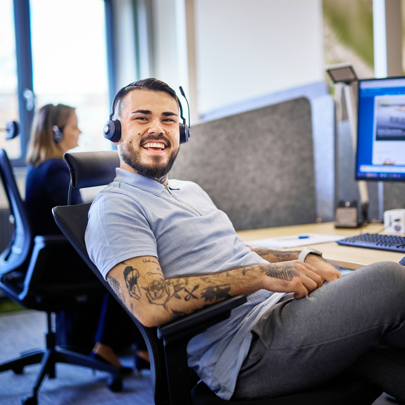 Junger Mann mit Headset und Tattoos lächelt entspannt am Schreibtisch in einem Büro mit Computer und Kollegin im Hintergrund