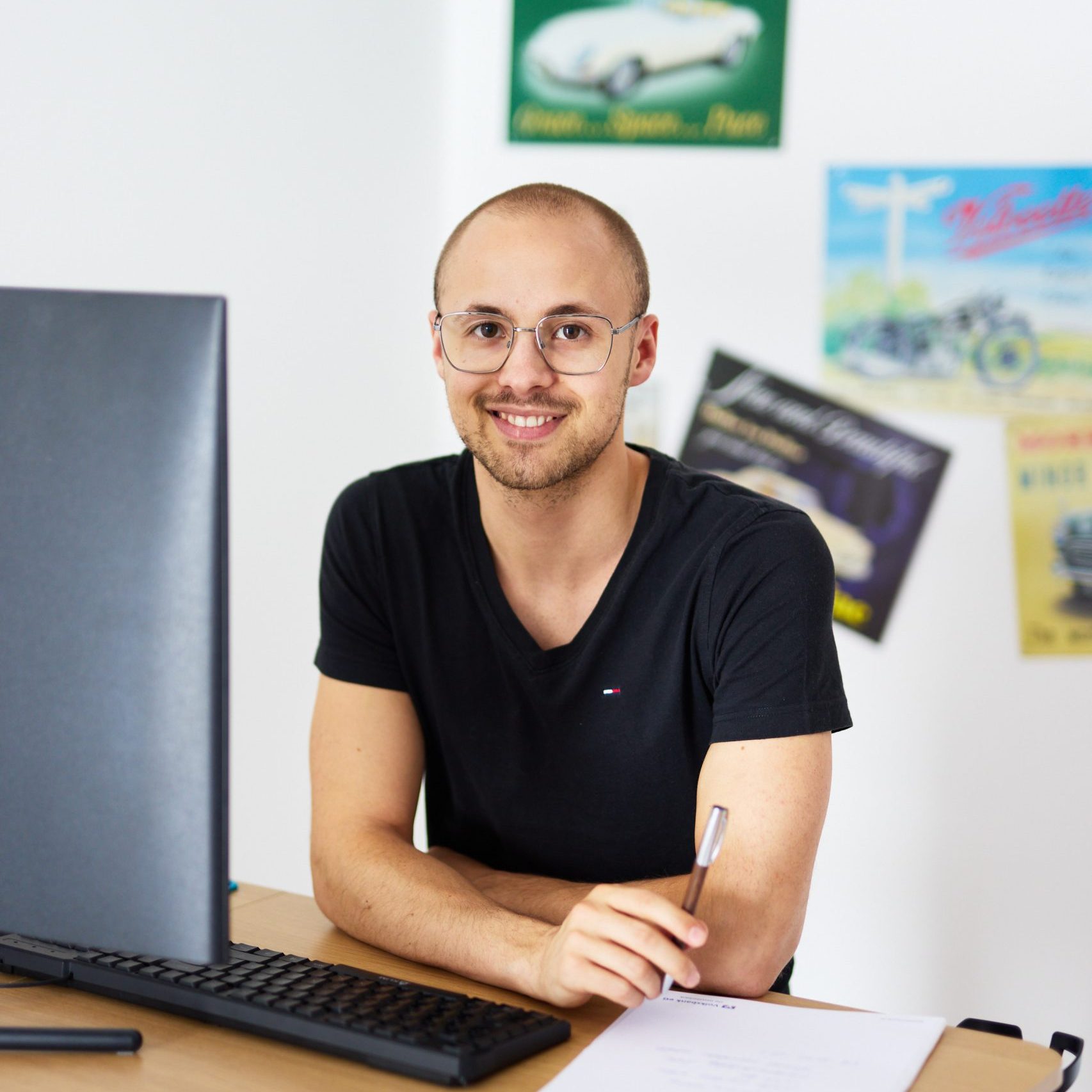 Junger Mann mit Brille und schwarzem T-Shirt sitzt lächelnd am Schreibtisch mit Tastatur und Stift in der Hand